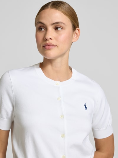 Kobiece model prezentuje Slim Fit Strickjacke aus Baumwoll-Mix od Polo Ralph Lauren, biały kolor, krój slim fit, krótki rękaw, okrągły dekolt, faktura dzianinowa, z przodu haftowane logo, jednolity wzór, zapięcie na guziki, materiał bawełniany, półzbliżenie na górną część ciała