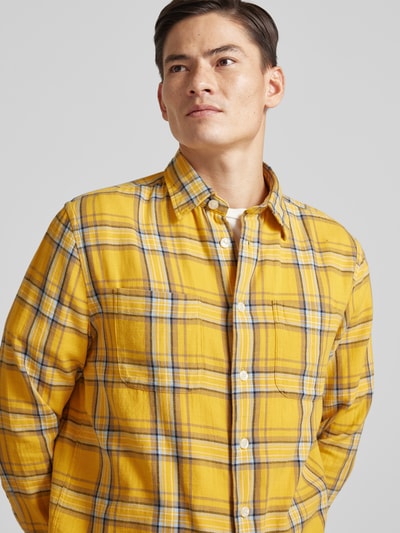 Męski model nosi Relaxed Fit Freizeithemd aus reiner Baumwolle od Tommy Hilfiger, kratka, długi rękaw, kołnierzyk kent, splot twill, kolor żółty, wyhaftowane logo, klasyczna listwa guzikowa, kieszenie na piersi; koszula casualowa z bawełny; półzbliżenie, widok z przodu.