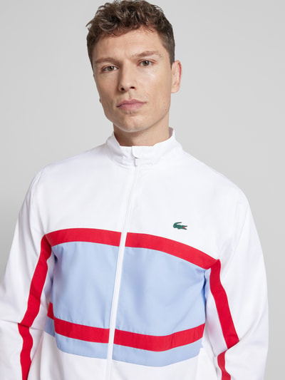 Männermodel trägt Regular Fit Trainingsanzug mit Kontraststreifen von Lacoste Sport, weiß mit roten und blauen Streifen, Nahaufnahme Oberkörper.