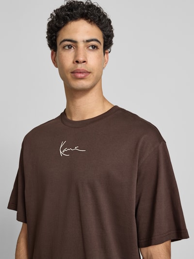 Männermodel trägt Regular Fit T-Shirt mit Logo-Stitching von KARL KANI in Braun, Rundhalsausschnitt, Kurzärmelig, Nahaufnahme Oberkörper, Rückenlänge bei Größe M: 74 cm.