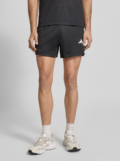 Mężczyzna nosi Straight Fit Shorts mit Logo-Print in Mesh-Optik von adidas Training, czarne, siateczkowa faktura, elastyczny pas, szerokość w rozmiarze M: 39 cm, długość wewnętrzna nogawki: 12 cm, materiał matowy, Ganzkörperansicht z przodu.