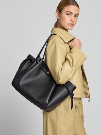 Frauenmodel trägt Handtasche mit Logo-Detail von Tory Burch in Schwarz mit Strukturmuster, Magnetverschluss, schmalen Schulterriemen, Ganzkörperansicht von vorne.