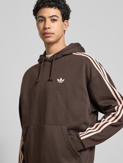 Männermodel trägt Oversized Hoodie mit Kapuze von adidas Originals in Braun mit rosa Streifen, Label-Stitching, Nahaufnahme Oberkörper