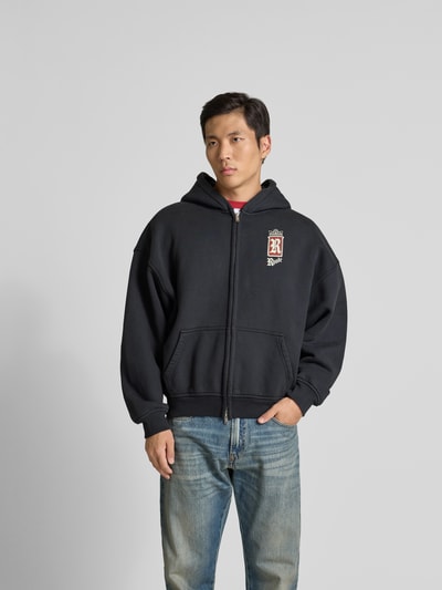 Männermodel trägt Oversized Sweatjacke aus Baumwolle von RHUDE in Schwarz mit Zweiwege-Reißverschluss und Känguru-Tasche, Ganzkörperansicht von vorne. Rückenlänge bei Größe M: 65 cm, Schulterbreite 60 cm, Ärmellänge 56 cm.