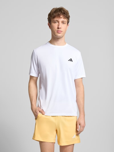 Mannenmodel draagt T-shirt met labelprint van adidas Training in wit met zwarte print, regular fit, ronde hals, ruglengte maat M 72 cm. Ganzlichaansicht van voren.