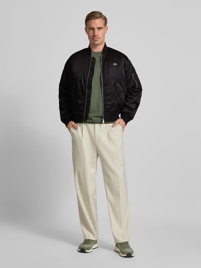 Männermodel trägt Regular Fit Bomberjacke aus Satin von Lacoste in Schwarz, unifarben, College-Kragen, Logo-Patch, Ganzkörperansicht von vorne.