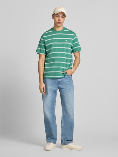 Männermodel trägt T-Shirt mit Logo-Stitching von Calvin Klein Jeans in Grün mit weißen Streifen aus Reine Baumwolle, Gerader-Schnitt, Rundhalsausschnitt, Rückenlänge bei Größe M 69 cm, Ganzkörperansicht von vorne.