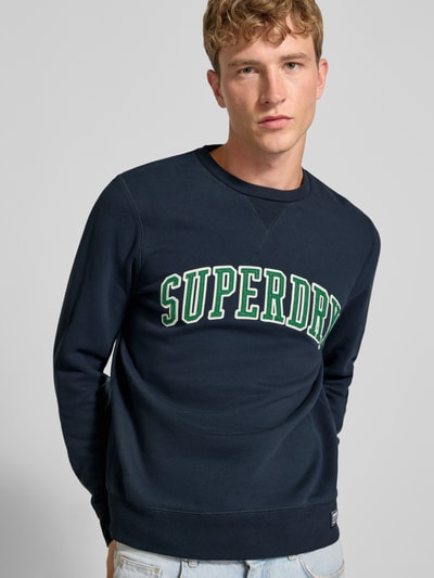 Männermodel trägt Regular Fit Sweatshirt aus Baumwoll-Mix mit Logo-Stickerei von Superdry, Dunkelblau, Rundhalsausschnitt, Ganzkörperansicht von vorne.