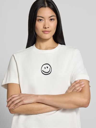 Kobiece model nosi Damski t-shirt Boxy Fit T-Shirt aus reiner Baumwolle model 'SARA' von VERO MODA, biały z czarnym uśmiechem z przodu, gładka faktura, zbliżenie na górną część ciała.