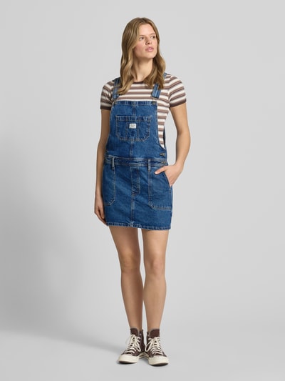 Frauenmodel trägt Jeanskleid mit Brusttasche und Label-Detail von Guess Jeans, Baumwoll-Elasthan-Mix, Gerader-Schnitt, Minikleid, Eingrifftaschen, Verstellbarer-Träger, Ganzkörperansicht von vorne.