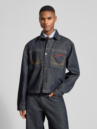 Männermodel trägt Relaxed Fit Jeansjacke mit Ziernähten von KARL KANI in dunklem Denim, Kontrastnähte, label-stitching, ganzkörperansicht von vorne