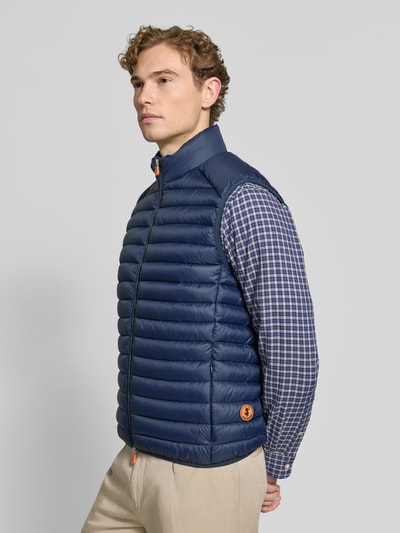 Männermodel trägt Steppweste mit Zweiwege-Reißverschluss Modell 'ADAM' von Save The Duck in Dunkelblau, Stehkragen, Ganzkörperansicht von vorne, Regular Fit, glatte gesteppte Textur, Rückenlänge 61 cm, Schulterbreite 45 cm, Brustweite 57 cm.
