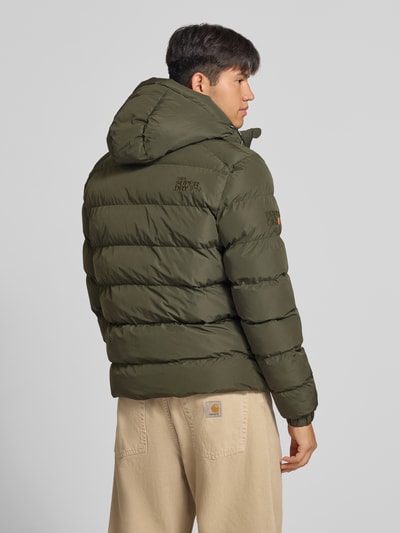 Männermodel trägt Steppjacke mit Label-Stitching und Kapuze von Superdry in Olivgrün, sichtbare Steppnähte, Ganzkörperansicht Rückansicht. Gerader Schnitt, Kapuze mit Kordelzug, Reißverschluss, seitliche Eingrifftaschen, elastische Ärmelabschlüsse, Länge 67 cm, Schulterbreite 48 cm, Brustweite 58 cm, Ärmellänge 72 cm.