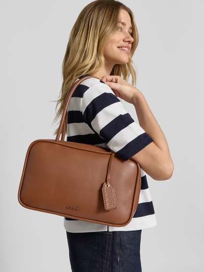 Frauenmodel trägt Henkeltasche in Lederoptik Modell 'JODI_DUFFLE' von HUGO, braun, Label-Applikation, Tragehenkel, Nahaufnahme Oberkörper.