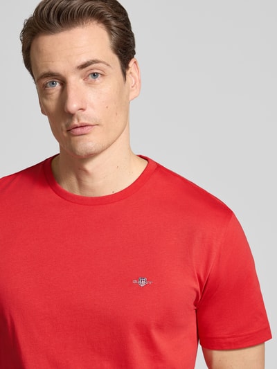 Męski model nosi Regular Fit T-Shirt mit Logo-Stitching od Gant z czystej bawełny, czerwony, krój regular fit, okrągły dekolt, długość tyłu w rozmiarze M: 68 cm, zbliżenie na górną część ciała z przodu.