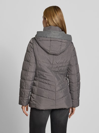 Frauenmodel trägt Jacke in Stepp-Optik mit Kapuze von FUCHS SCHMITT, taliowany krój, kaptur, z ociepleniem, wpuszczane kieszenie z zamkiem błyskawicznym, Długość tyłu w rozmiarze 36: 74 cm, Szerokość ramion w rozmiarze 36: 40 cm, Szerokość klatki piersiowej w rozmiarze 36: 48 cm, Długość rękawów w rozmiarze 36: 64 cm, szare odcienie, pikowana tekstura, Ganzkörperansicht Rückansicht.