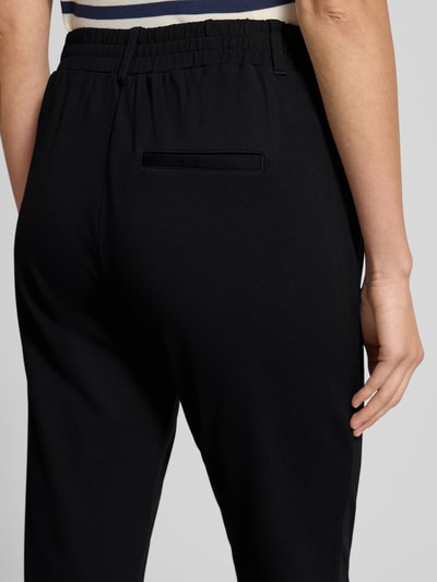 Kobiece modelka nosi Easy Pants mit Bundfalten od TOM TAILOR, czarny kolor, mieszanka wiskozy z dodatkiem streczu, ozdobna listwa z tyłu, elastyczny pas, tunel, zakładki w pasie, wpuszczane kieszenie, zwężane nogawki, widok całych spodni z tyłu.