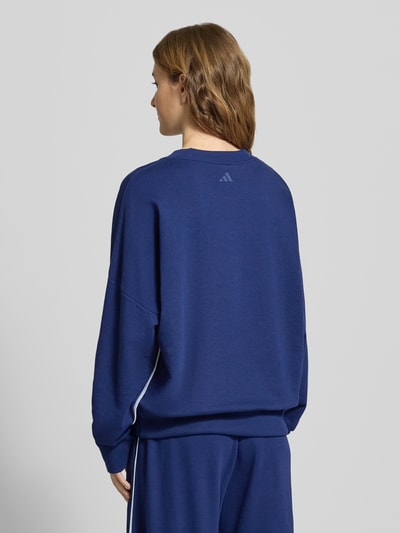 Kobiece modelka nosi Sweatshirt mit Label-Stitching od adidas Sportswear, bawełniana mieszanka z domieszką rayonu, luźny krój, ściągacze, okrągły dekolt, haftowane logo, granatowy kolor, tekstura miękka, widok całej sylwetki z tyłu.