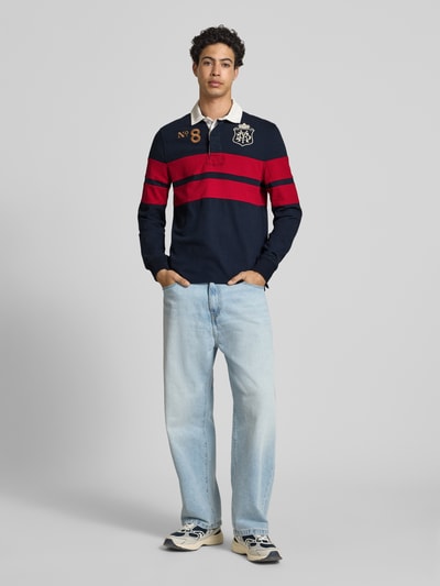 Männermodel trägt Regular Fit Rugby Shirt mit Logo- und Motiv-Stitching von Superdry in Dunkelblau mit roten Streifen, Baumwolle, Ganzkörperansicht von vorne.