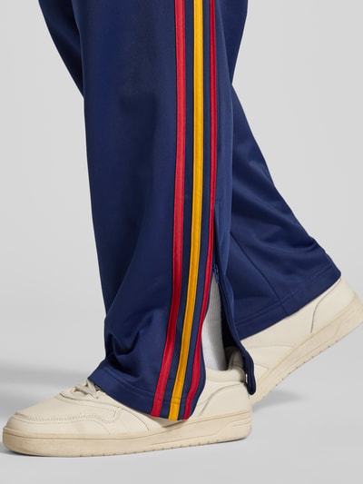 Männermodel trägt adidas Originals Regular Fit Trackpants aus recyceltem Material mit Logo-Stitching und mehrfarbigen Streifen, Detailaufnahme.