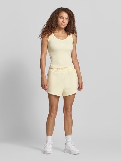 Frauenmodel trägt High Waist Sweatshorts mit Logo-Stitching Modell 'WILUNA' von Pegador aus Baumwoll-Mix, sanftes Gelb, elastischer Bund, Struktur glatt, Bundweite Größe S 30 cm, Innenbeinlänge 8 cm, Ganzkörperansicht von vorne.