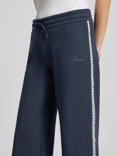 Kobieta nosi spodnie dresowe 'Wide Fit Sweatpants aus Baumwoll-Mix' od REVIEW, granatowe, mieszanka bawełny, bordiura, wyhaftowane logo, długość wewnętrzna nogawki w rozmiarze S: 44 cm, zbliżenie na górną część nogawek z przodu.