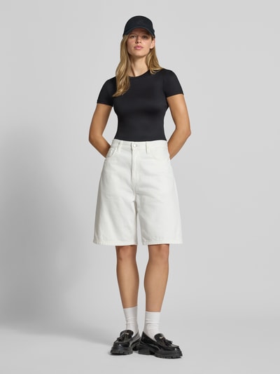 Frauenmodel trägt schwarzes Slim Fit T-Shirt mit Rundhals Modell 'LULA' von PIECES, kombiniert mit weißer Bermuda, Ganzkörperansicht von vorne.