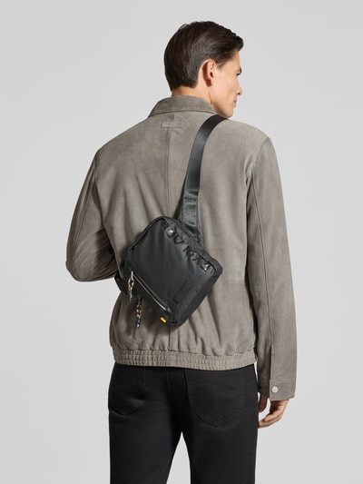 Männermodel trägt Crossbody Bag mit Statement-Print Modell 'EXPLORE' von camel active, schwarze Farbe, Reißverschlussfächer, Ganzkörperansicht Rückansicht.