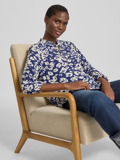 Modelka kobieca ma na sobie Regular Fit Bluse aus Viskose mit 3/4-Arm marki Marc Cain, wiskoza, wzór na całej powierzchni, stójka, wiązanie, rękaw o dł. 3/4, długość tyłu w rozmiarze 36: 63 cm, długość rękawów w rozmiarze 36: 50 cm, kolory granatowy i kremowy, widoczne miękkie, gładkie tekstury, ujęcie: zdjęcie całej sylwetki z przodu.