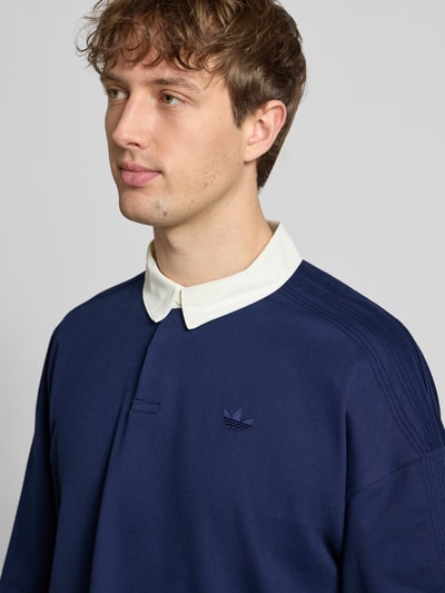 Männermodel trägt Regular Fit T-Shirt aus reiner Baumwolle von adidas Originals, navyblau mit weißem Polo-Kragen, Logo-Stitching, Nahaufnahme Oberkörper.