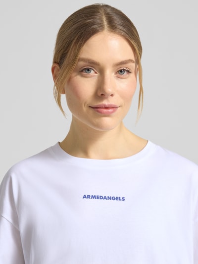 Modelka kobieca nosi Oversized Langarmshirt aus reiner Baumwolle Modell 'FIETAA SMILE' od Armedangels, biała gładka bawełna, logo haftowane, detal z przodu, krój oversize, dekolt okrągły, długie rękawy, drobny ściągacz, widok z przodu, zbliżenie na górną część ciała.