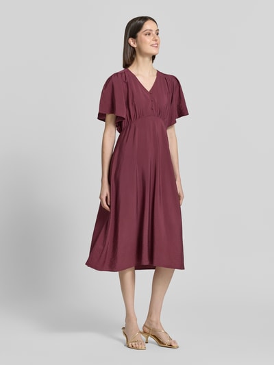 Frauenmodel trägt Midikleid aus Viskose-Mix mit V-Ausschnitt Modell 'Aniva' von ICHI, Tailliert, Kurzärmelig, Knopfleiste, Ganzkörperansicht von vorne. Rückenlänge bei Größe 36: 116 cm, Brustweite: 34 cm, Ärmellänge: 26 cm. Glatte, weinrote Textur, lockere Passform/Schnitt:.