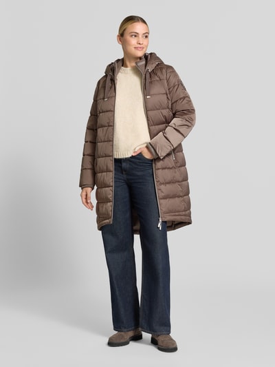 Vrouwenmodel draagt Regular Fit lange jas in stiksels van monari, capuchon met tunnelkoord, opstaande kraag, steekzakken met rits, tweewegritssluiting, bruine kleur, zachte textuur, ruglengte maat 36: 100 cm, schouderbreedte: 43 cm, borstwijdte: 49 cm, mouwlengte: 65 cm, full body vooraanzicht.