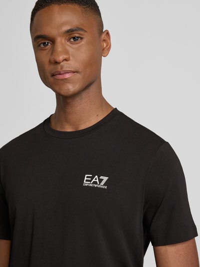 Männermodel trägt schwarzes T-Shirt mit Label-Print von EA7 Emporio Armani, Slim Fit, Baumwoll-Elasthan-Mix, Nahaufnahme Oberkörper.