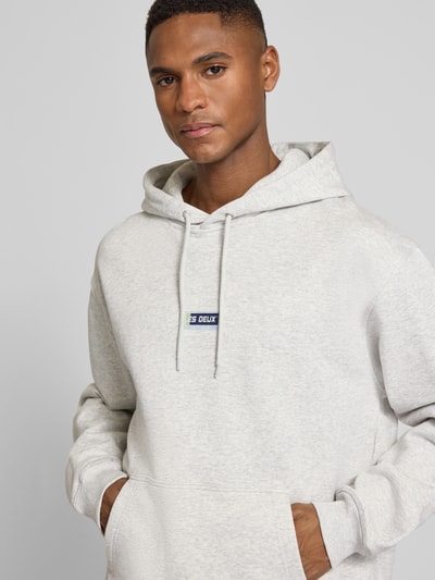 Männermodel trägt Hoodie mit Logo-Print Modell 'Chase' von Les Deux aus Reine Baumwolle im Loose-Fit mit Kapuze, Kordelzug, Känguru-Tasche, elastische Abschlüsse, hellgraue Farbe, melierte Textur, Nahaufnahme Oberkörper.
