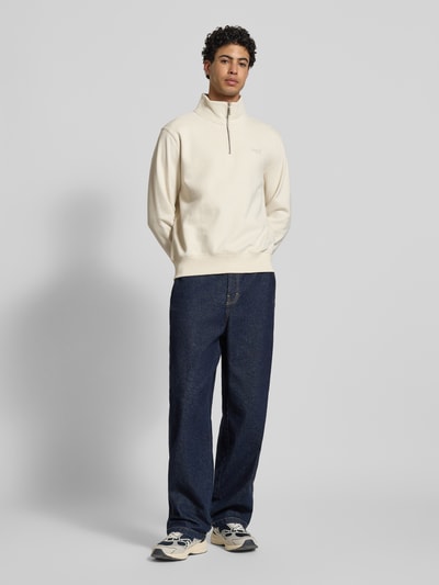 Männermodel trägt Regular Fit Sweatshirt mit Logo-Stitching von Superdry in Creme, Baumwoll-Mix, Stehkragen mit Reißverschluss, elastische Abschlüsse, Rückenlänge 65 cm, Ärmellänge 63 cm, Ganzkörperansicht von vorne.