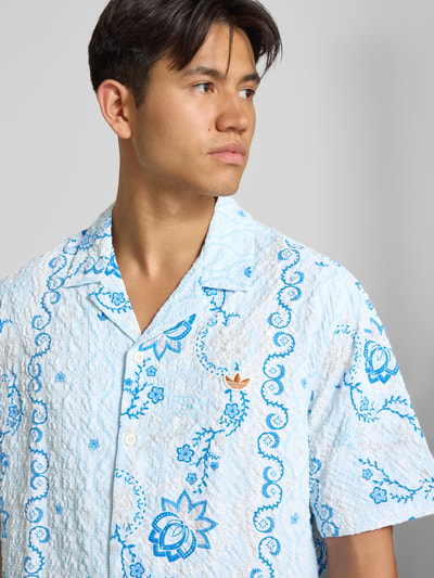 Männermodel trägt Relaxed Fit Freizeithemd aus Seersucker mit Logo-Stitching von adidas Originals, weiß-blaues Muster, Nahaufnahme Oberkörper.