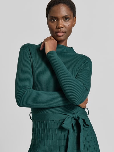 Kobiece model prezentuje Knielanges Strickkleid mit Bindegürtel od Apricot, Mieszanka wiskozy, Linia a, Długość do kolan, Stójka, wiązany pasek, Długość tyłu w rozmiarze S: 103 cm, Szerokość w rozmiarze S: 38 cm, Długość rękawów w rozmiarze S: 59 cm, zielony, tkanina o prążkowanej oraz plecionej fakturze, zbliżenie na górną część ciała.