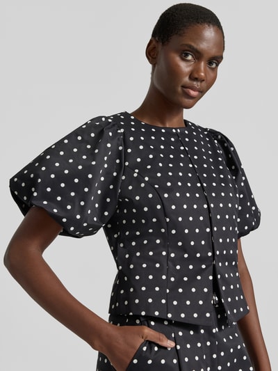 Frauenmodel trägt Slim Fit Bluse im Allover-Look Modell 'ANELA' von Neo Noir, schwarzes Weißpunktmuster, Rundhalsausschnitt, Puffärmel, Hakenverschluss, Ganzkörperansicht von vorne.