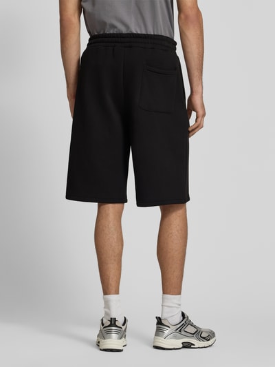 Männermodel trägt Sweatshorts mit Logo-Print von REVIEW in Schwarz, Material-Mix mit Baumwoll-Anteil, Loose-Fit, elastischer Bund, Ganzkörperansicht Rückansicht.