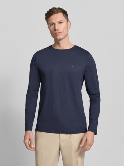 Mężczyzna nosi bluzkę z długim rękawem z okrągłym dekoltem i logo Longsleeve mit Rundhalsausschnitt und Logo od Fynch-Hatton, bawełna, krój regular fit, jednokolorowy, długość tyłu 70 cm, rozmiar M, widok całej sylwetki z przodu.