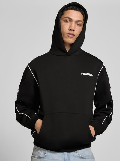 Męski model nosi Hoodie mit Logo und Känguru-Tasche od REVIEW, czarny bawełniany krój regular fit, logo, kieszeń kangurka, długość tyłu w rozmiarze M: 71 cm, długość rękawa w rozmiarze M: 67 cm, widok całej sylwetki z przodu.
