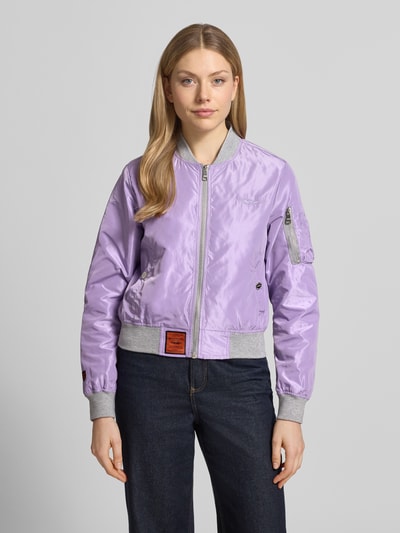 Frauenmodel trägt Bomberjacke mit Label-Patch und Reißverschlusstasche am Ärmel von Original Bombers in Lila mit grauen Bündchen, Lockerer-Schnitt, seitliche Eingrifftaschen mit Druckknopfverschluss, Stehkragen, Reißverschluss, Logo-Stitching, Maße: Rückenlänge 53 cm, Schulterbreite 37 cm, Brustweite 48 cm, Ärmellänge 63 cm, Stoff glänzend, Ganzkörperansicht von vorne.