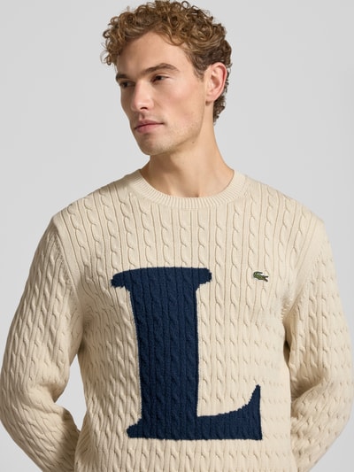 Männermodel trägt Regular Fit Strickpullover aus Baumwoll-Mix von Lacoste in Creme mit blauem L und Logo, Nahaufnahme Oberkörper. Herren Strickjacke von Lacoste.