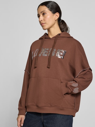 Frauenmodel trägt Oversized Hoodie aus reiner Baumwolle Modell 'L(I)EBE' von miss goodlife in Braun mit Pailletten- und Strasssteinbesatz, Känguru-Tasche, gerippte Abschlüsse, Nahaufnahme Oberkörper.