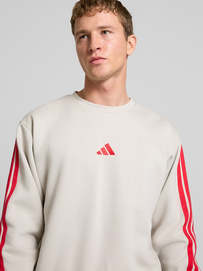 Männermodel trägt Sweatshirt mit Label-Stitching von adidas Sportswear in Beige mit roten Streifen, Baumwoll-Elasthan-Mix, Nahaufnahme Oberkörper.