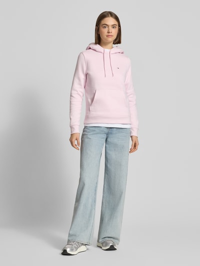 Modelka kobieca ma na sobie Regular Fit Hoodie aus Baumwoll-Mix od Tommy Jeans, różowa bawełniana bluza z kapturem, gładka faktura, detal z logo, długie rękawy, ściągacze, materiał sweat, krój regular fit, widok całej sylwetki z przodu.