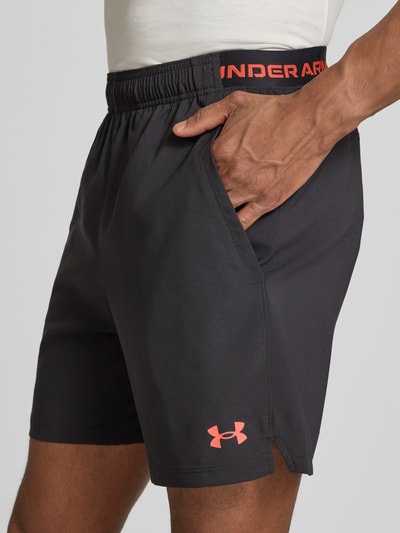 Mężczyzna nosi Shorts mit Logo und Eingrifftaschen von Under Armour, czarny kolor, krój regular fit, logo, elastyczny pas, szerokość w rozmiarze M: 38 cm, zbliżenie na górną część ciała.