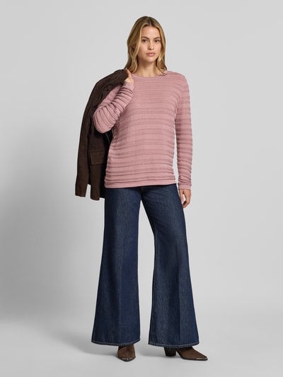 Frauenmodel trägt Regular Fit Pullover aus reiner Baumwolle in Strick-Optik von Christian Berg Woman in Rosa, semitransparent, feine horizontale Linien, kombiniert mit dunkelblauer Schlagjeans, Ganzkörperansicht von vorne.