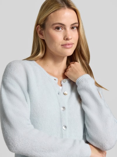 Frauenmodel trägt Relaxed Fit Strickjacke aus Woll-Mix mit Leinen-Anteil von American Vintage in Hellblau, weiche Textur, Nahaufnahme Oberkörper. Damen Strickjacke mit gerippten Bündchen, durchgehender Knopfleiste, langärmlig, Rückenlänge 52 cm, Ärmellänge 56 cm.